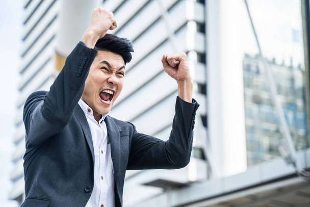 就職面接試験の結果を知って、アジアの若いビジネスマンは幸せな気持ちになります。市内で幸せを手と腕を上げている男は、オフィスの仕事を得るために喜んでいます. - 写真・画像