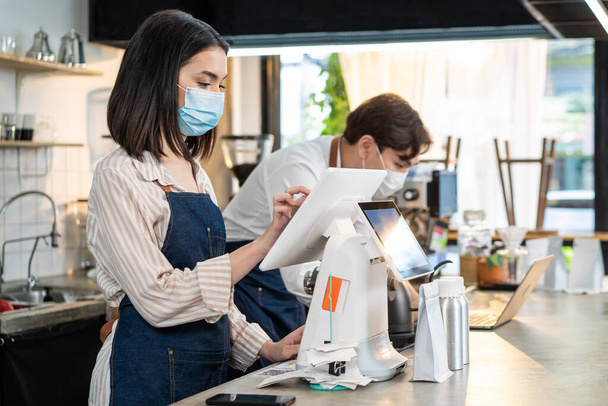 Asian cafe business wear mask, use laptop check food order and payment. Young attractive beautiful waiter use touch screen on counter bar to prepare food for pick up, takeaway during Covid 19 pandemic - Foto, imagen