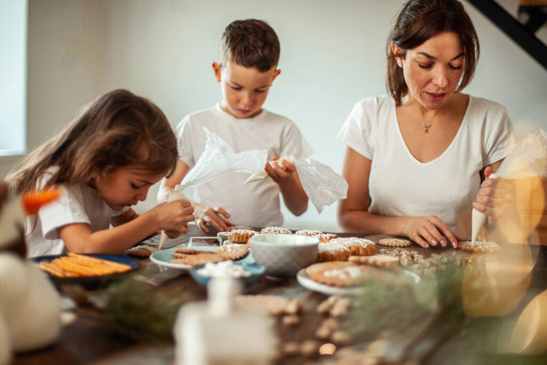 ママと子供たちは自宅でクリスマスジンジャーブレッドを飾る。男の子と女の子がクッキーに砂糖アイシングでコーネットでペイントします。新年の装飾、クリスマスツリーの枝. - 写真・画像