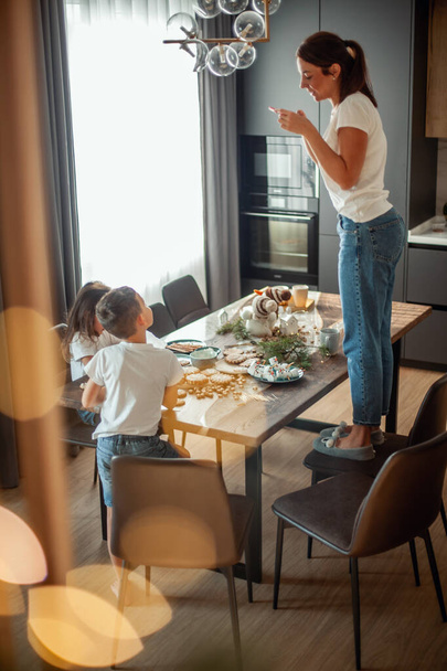 Mom takes pictures of topview as daughter and son decorate gingerbread with sugar icing. Stylish home dining and kitchen. - Φωτογραφία, εικόνα