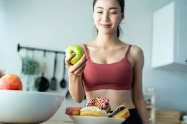 Asian attractive active woman pick up a green apple in kitchen at home. Beautiful sport girl in sportswear desire to chose fruit and ignore sweet donut enjoy eat healthy food for health after exercise - 写真・画像