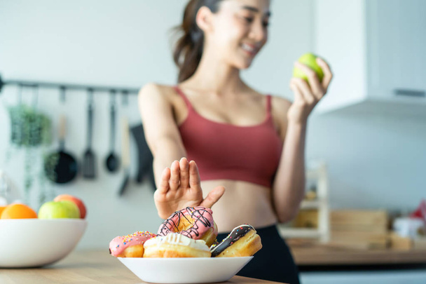 Ασιατική ελκυστική ενεργή γυναίκα πάρει ένα πράσινο μήλο στην κουζίνα στο σπίτι. Όμορφο κορίτσι του αθλητισμού στα αθλητικά επιθυμία να επιλέξει φρούτα και να αγνοήσει γλυκό ντόνατ απολαύσετε τρώνε υγιεινά τρόφιμα για την υγεία μετά την άσκηση - Φωτογραφία, εικόνα