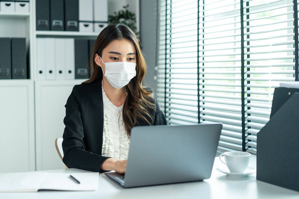 Azjatka w garniturze nosi maskę i pracuje w miejscu pracy. Atrakcyjna pracownica biurowa siedzi na stole, korzysta z laptopa, aby omówić plan firmy podczas pandemii Covid19 - Zdjęcie, obraz