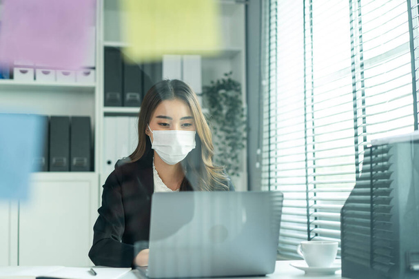 Asian businesswoman in formal suit wear facemask and work at workplace. Attractive female employee office worker sit on table, use laptop computer to discuss about company plan during Covid19 pandemic - Foto, imagen