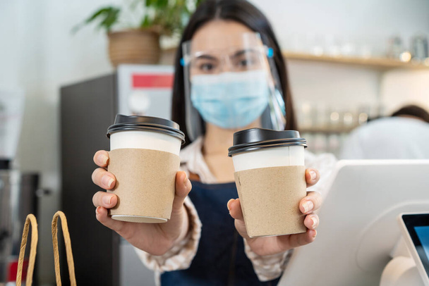 Portrait of restaurant waiter wear mask handing cup of coffee in cafe. Happy Attractive beautiful woman worker work in coffeehouse and holding beverages cups with two hands due to Covid-19 pandemic. - Фото, изображение