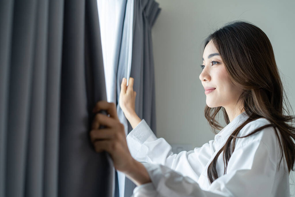 Asian beautiful woman approaches a window and open curtains in morning. Young attractive girl in white pajamas feel fresh and happy to look outside and sunrise after wake up early in bedroom at home. - Fotografie, Obrázek