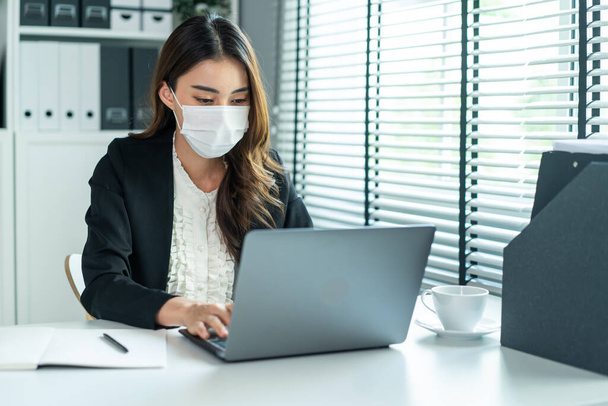 Asian businesswoman in formal suit wear facemask and work at workplace. Attractive female employee office worker sit on table, use laptop computer to discuss about company plan during Covid19 pandemic - Zdjęcie, obraz