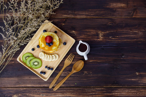 plato de madera de lujo sabroso panqueque saludable cubierta por fresa, arándano, y el corte de plátano y kiwi servido en la mesa de comedor marrón con un pequeño frasco de jarabe dulce, tenedor, y cuchara - Foto, Imagen