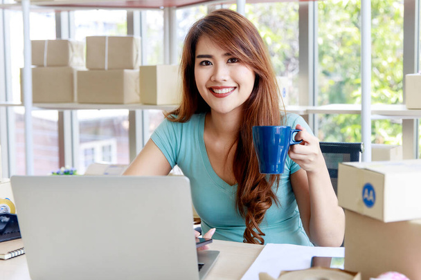 hermosa empleada sonríe felizmente como ella va a tener un descanso con la taza de café en el escritorio de trabajo que rodea por cajas de embalaje en la oficina del negocio de servicios de entrega - Foto, imagen