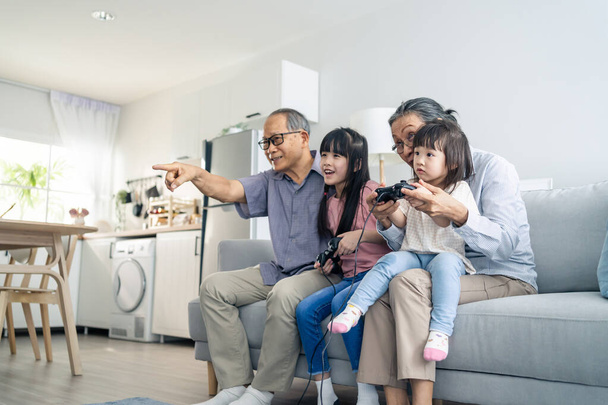 Ασιατική ευτυχισμένη οικογένεια διαμονή στο σπίτι παππούς παιχνίδι με το κοριτσάκι. Αγαπώντας ηλικιωμένα ηλικιωμένα ζευγάρια παίζουν βιντεοπαιχνίδια μπροστά στην τηλεόραση με μικρά παιδιά εγγονή στο σαλόνι μαζί. - Φωτογραφία, εικόνα
