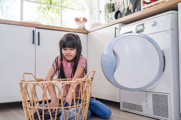 Asian young little kid put dirty clothes into washing machine in house. Lovely girl child sit on floor, feel happy to help family and loads laundry in washer appliance at home. Domestic-House keeping. - Photo, image