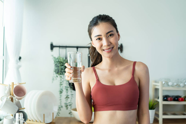 Portrét atraktivní asijské ženy nalévat vodu do skla v kuchyni. Mladý žíznivý krásný sport dívka ve sportovním oblečení držet čisté minerální přírodní v šálku po cvičení cvičení pro zdravotní péči v domě. - Fotografie, Obrázek