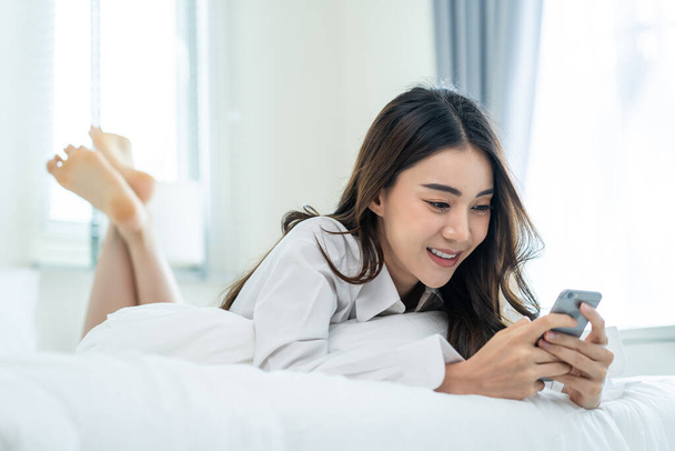 Asian attractive woman use mobile phone chat on bed at home in morning. Happy casual beautiful female lying down in bedroom enjoy holiday weekend, use smartphone communicate with technology in house. - Photo, image