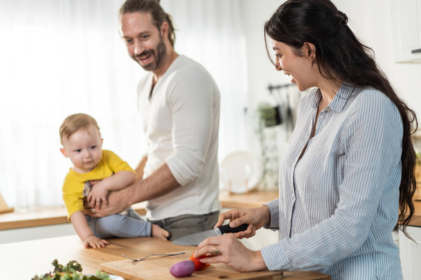 Beyaz bir aile, anne mutfakta baba ve bebekle yemek pişiriyor. Sevgi dolu baba kucağında küçük bir çocuk taşır, annemle evde vakit geçirirken eğlenir. Etkinlik ilişkisi kavramı. - Fotoğraf, Görsel