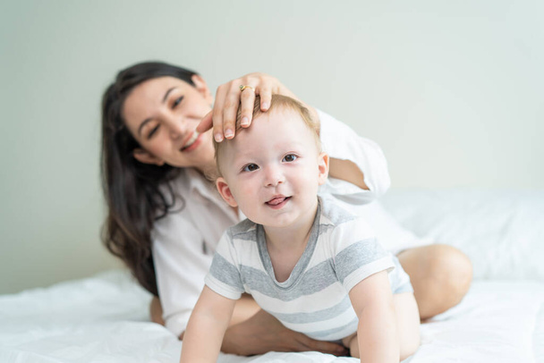 Portrait of Caucasian happy family smiling, look at camera in bedroom. Young attractive woman parents, mother in pajamas laying on bed with little baby boy child enjoy morning wake up activity at home - Foto, Bild
