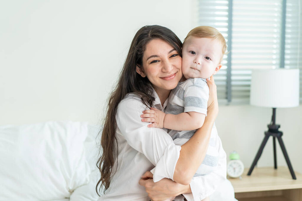 Portrait of Caucasian happy family smiling, look at camera in bedroom. Young attractive woman parents, mother in pajamas sit on bed with little baby boy child enjoy morning wake up activity in house. - Photo, Image