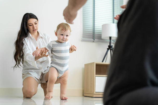 Caucasian young cute baby boy child learn to walk with parent in house. Happy family mother support little toddler son taking first step walk on floor to father to develop skill in living room at home - Photo, Image