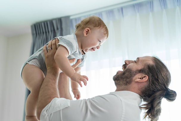 Caucasian loving father tosses cute baby boy child up in living room. Happy family, attractive caring young daddy carry his toddler son on hands with happiness in house. Activity relationship at home. - Foto, afbeelding