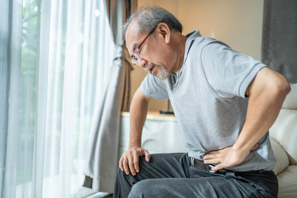 Aziatische oudere man voelt zich thuis vreselijk last van buikpijn. Eenzame gestresste oudere volwassen man zitten op de bank met een pijnlijk gezicht aanraken van de buik. Begrip ziektekostenverzekering. - Foto, afbeelding