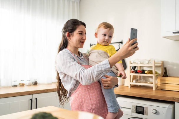 Caucasian mother holding little baby and video call online with family. Attractive beautiful mom using mobile cellphone and chat with husband while carrying young toddler kid in living room in house. - Zdjęcie, obraz