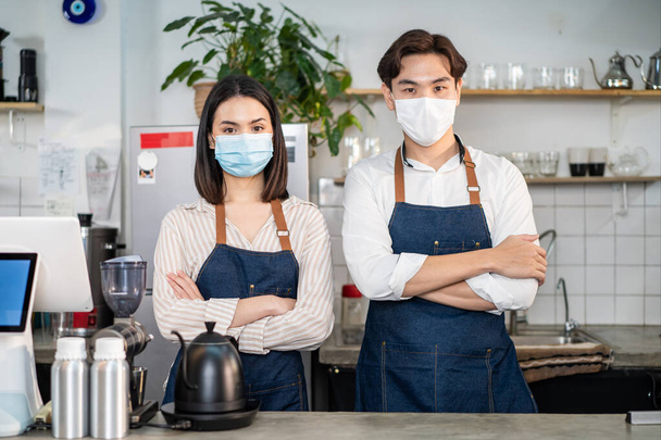 Portrait of attractive Asian waiter and waitress at restaurant cafe. Barista business couple in apron wearing protective mask , stand with confident and crossing arm due to Covid-19 at coffee shop. - Photo, image