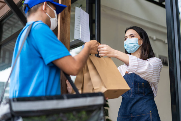 アジアの食品配達人は、 Covid-19パンデミックのために保護マスクを着用し、コーヒーショップのレストラン労働者に電話でオンライン注文を示しています。ウエートレスは新しい通常のライフスタイルのための郵便配達人に持ち帰りバッグを与える. - 写真・画像