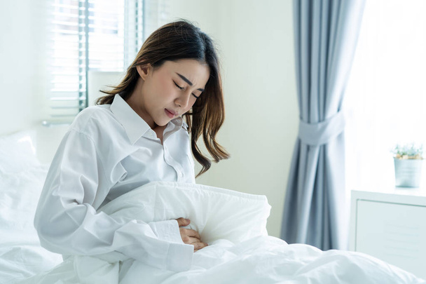 Ázsiai gyönyörű beteg lány pizsamában, aki a hálószobában alszik. Vonzó fiatal nő rosszul és fájdalmasan érzi magát, miután felébredt az ágyban, fáj a gyomra vagy menstruál, majd reggel hascsikarás.. - Fotó, kép