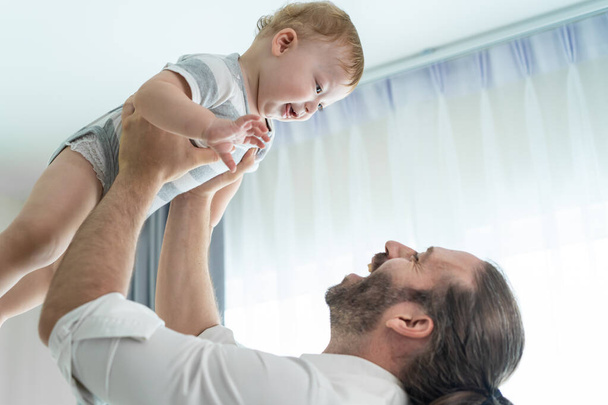 Caucasian loving father tosses cute baby boy child up in living room. Happy family, attractive caring young daddy carry his toddler son on hands with happiness in house. Activity relationship at home. - Valokuva, kuva