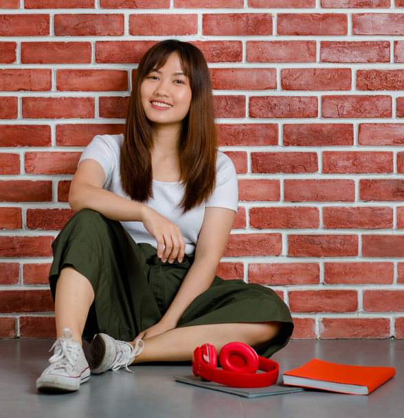Teljes hossza fiatal vonzó ázsiai nő fehér pólóban és zöld szoknya ül a padlón könyvek és piros fejhallgató a padlón ellen narancssárga tégla fal háttér. - Fotó, kép