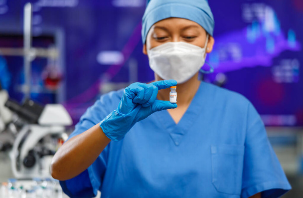 Ärztinnen mit Hygiene-Gesichtsmaske und bedeckter Kleidung bleiben im Laborraum und halten eine Flasche Covid-19 Impfstoff mit Laborwerkzeugen im Hintergrund. Selektiver Fokus - Foto, Bild