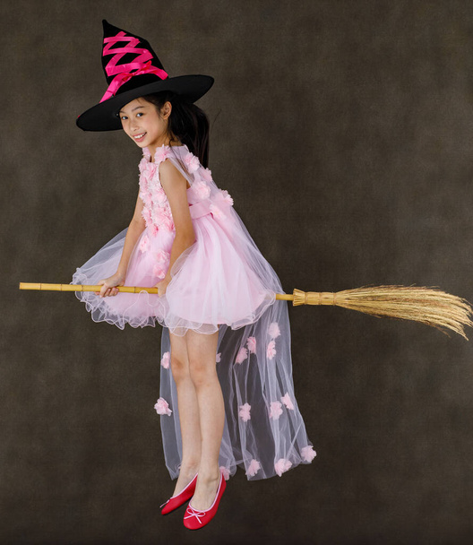 Portrait studio shot of little cute Asian kid in pink witch dress costume with black high hat look at camera posing flying gesture riding magic witchcraft broomstick on Halloween traditional festival. - Foto, Imagem