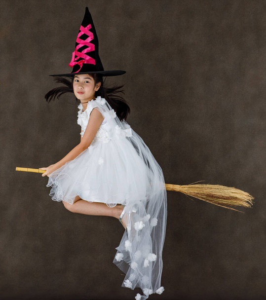 Portrait studio shot of little cute Asian kid in pink witch dress costume with black high hat look at camera posing flying gesture riding magic witchcraft broomstick on Halloween traditional festival. - Foto, Imagem