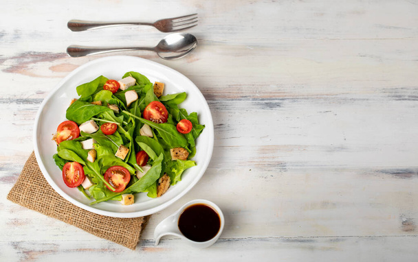 Fresh spinach salad with tomato, small pieces of herb roasted chicken in white ceramic dish on brown sack cloth, together with balsamic vinegar and metal utensil on white wooded table. - Photo, Image