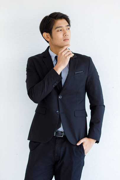Portraitaufnahme von asiatischen jungen gut aussehenden glücklichen Intelligenz erfolgreichen Geschäftsmann in schwarzen formalen Anzug stehen halten Hand berühren Kinn grübeln und lächeln allein vor weißem Wandhintergrund. - Foto, Bild