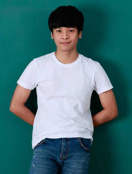 Entzückter junger asiatischer Mann in weißem T-Shirt und Jeans steht auf grünem Hintergrund im Studio und blickt in die Kamera - Foto, Bild