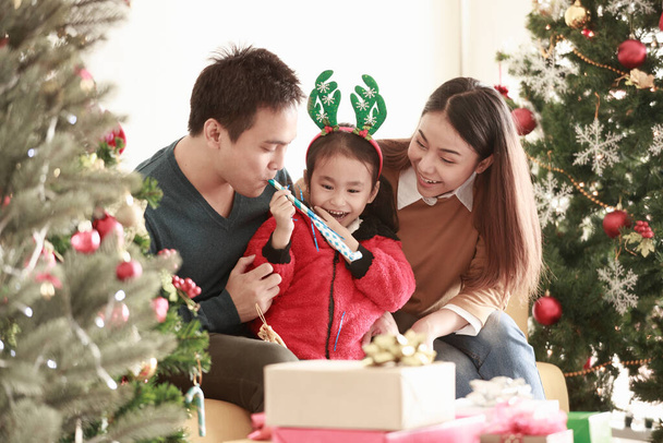 Asyalı gülümseyen kız mutlu bir şekilde anne ve babayla alay etti. Babam, annem ve Noel Baba şapkalı bebeğim Noel ağacının yanındaki kanepede oturuyorlar. Konsept Tease Love Cozy Gülümse sevimli - Fotoğraf, Görsel