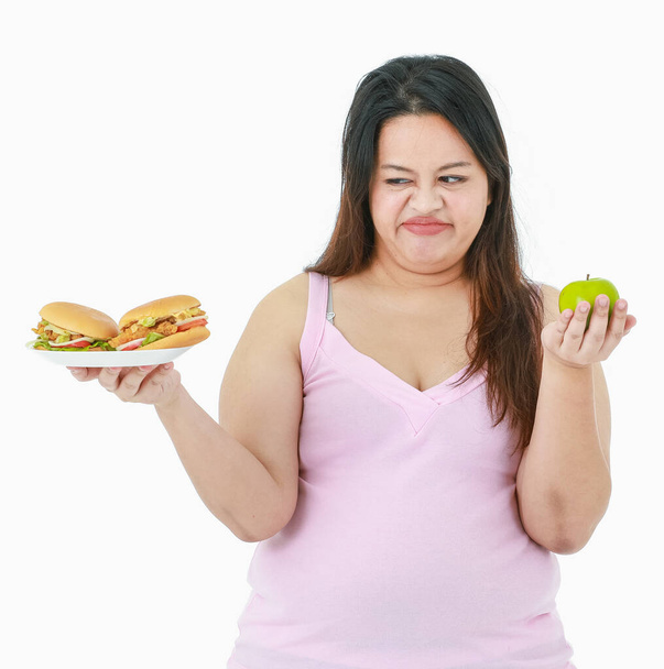アジアの濃縮ストレスの孤立したスタジオショット大きな脂肪太りすぎの女の子は手で健康的な有機緑のリンゴを食べることを決定し、嫌い前に不健康なチーズバーガーが好きではありません白い背景. - 写真・画像