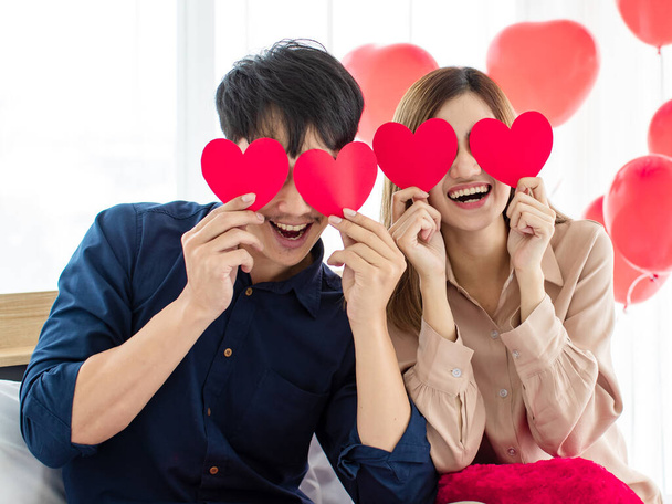 Juguetones asiáticos amantes del hombre y la mujer sonriendo y sosteniendo corazones de papel cerca de los ojos durante la celebración del Día de San Valentín. concepto de amor feliz pareja. - Foto, Imagen