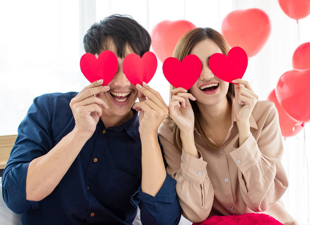 Játékos ázsiai férfi és nő szerelmeseinek mosolygós és tartó papír szív közel szemek alatt Szent Valentin-napi ünnepség. Boldog pár szerelem koncepció. - Fotó, kép