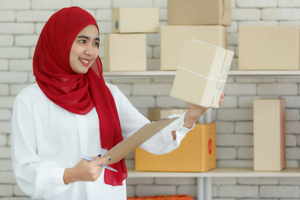 Portrait shot of cute smiling young teenage Muslim woman wearing red hijab standing while holding postage boxes and order checklist to double-check again before delivery. Concept of careful work - Photo, Image