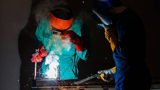 Un trabajador de la fábrica con un mono mecánico verde y casco de seguridad soldadura metalurgia en la noche en una fábrica, mientras que su colega mirando una chispa de soldadura. - Foto, imagen