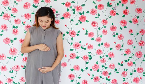 Porträt einer jungen schwangeren Asiatin, die lächelt und ihren Bauch berührt, während sie in eine Kamera über weißem Rosenmuster blickt. Gesundes und glückliches Damenkonzept. - Foto, Bild