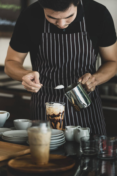 Fiatal ázsiai barista férfi fekete-fehér kötényben, kanállal, hogy szándékosan tejhabos kávét töltsön fel a pultnál az edények, üvegek, poharak által körülvett lágyság és lágyság érdekében. - Fotó, kép
