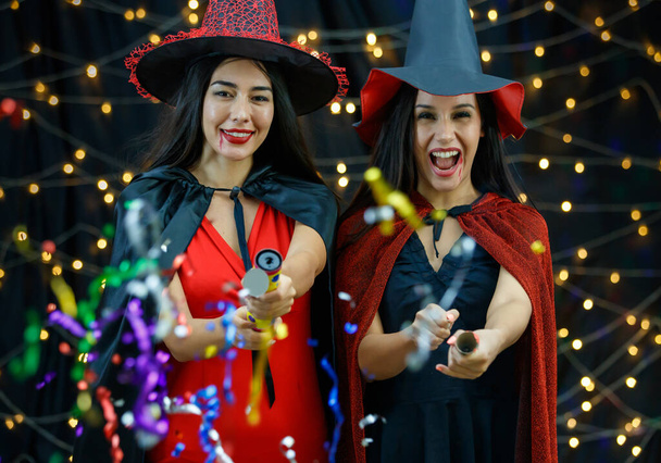 ハロウィンパーティー中にカメラに向かって蛇行と楽しいと爆発クラッカーを持っているスタイリッシュな魔女の衣装や帽子で楽しい若い女性の友人 - 写真・画像
