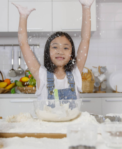 A little cute Asian girl, 7 years old tries to mix flour for bread baking during learning how to make a bakery in a modern kitchen. She spread flour into the air with fun and happiness. - Photo, Image