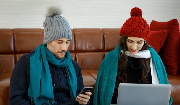 Nowoczesny styl życia kaukaski chłopak i dziewczyna kochanek para w wełnianej kurtce sweter dzianiny kapelusz i szalik siedzieć na podłodze oprzeć się na kanapie skupić się na osobistym urządzeniu cyfrowym smartfon i laptop. - Zdjęcie, obraz