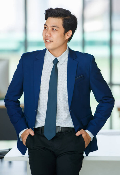Wesoły młody i przystojny azjatycki biznesmen relaks pozowanie stojąc ze szczęśliwym i pozytywnym działaniem w biurze miejsca pracy. - Zdjęcie, obraz
