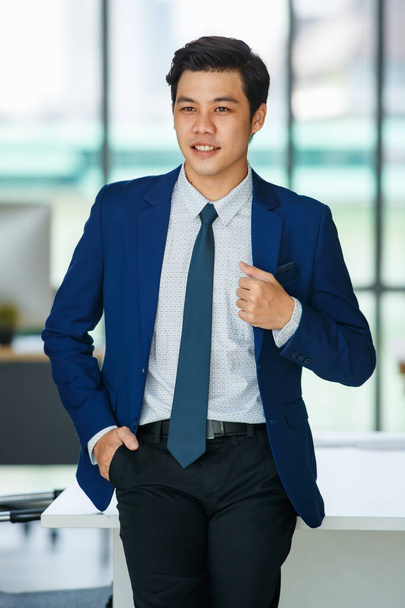 Веселый молодой и красивый азиатский бизнесмен расслабляется позируя, стоя со счастливым и позитивным действием в офисе на рабочем месте. - Фото, изображение