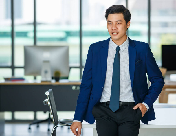 Vidám fiatal és jóképű ázsiai üzletember pihenni pózol, miközben áll a boldog és pozitív fellépés a munkahelyi irodában. - Fotó, kép