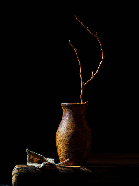 Otoño marchitándose minimalista Bodegón con rama marchita en jarrón de arcilla y hoja seca sobre mesa de madera Vertical, baja llave, vista lateral. Bodegón holandés - Foto, Imagen